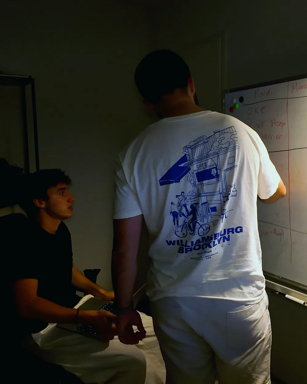 Nicolas écrit au tableau blanc, Paco assis à côté, au bureau le soir